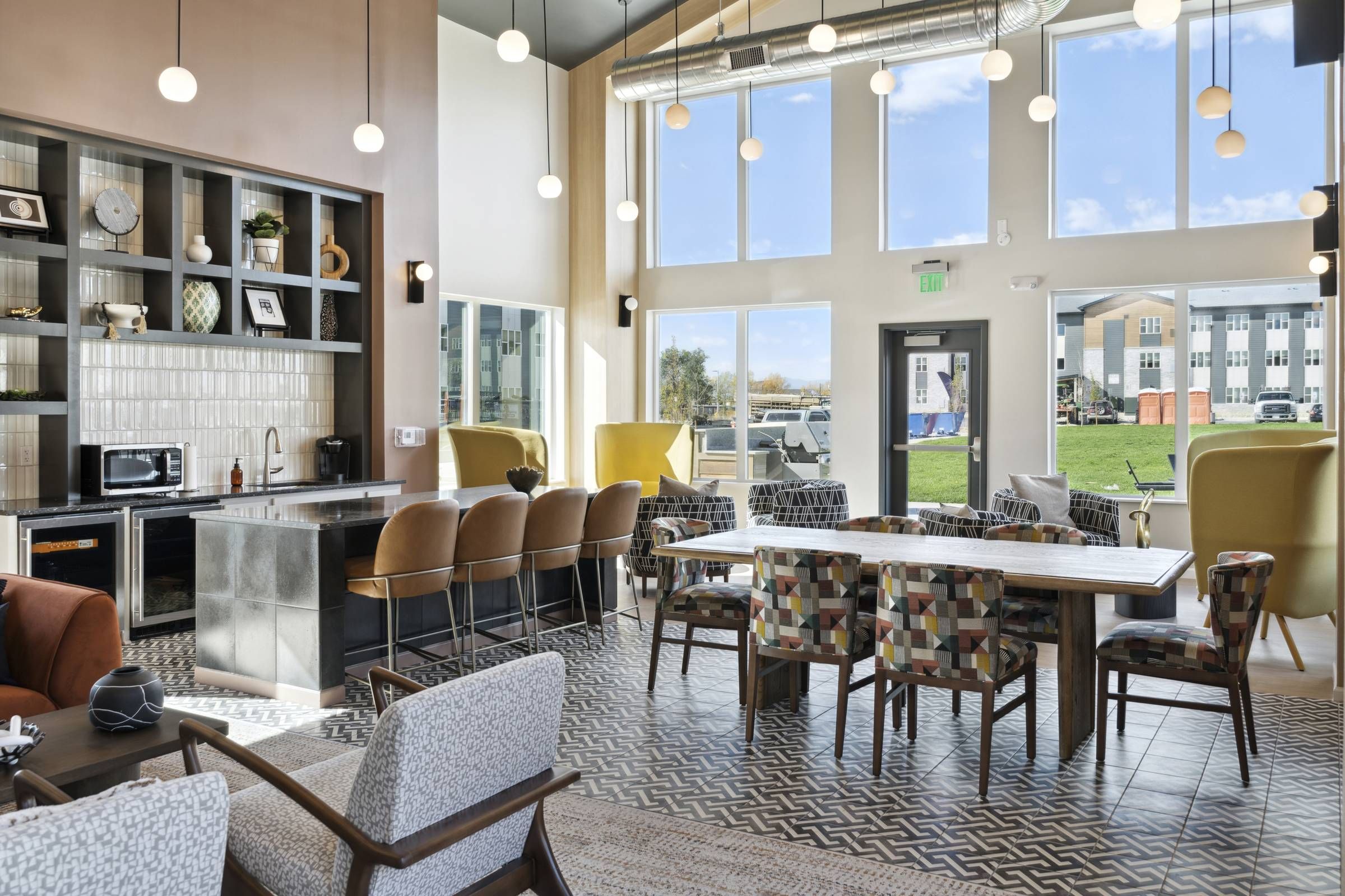 Modern communal lounge with patterned chairs, bar seating, tall windows, and bright natural light.